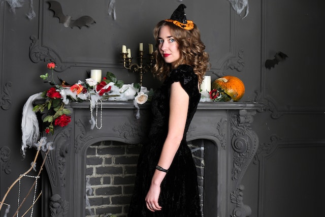 A girl dressed as a witch stands next to the fireplace celebrating Halloween in Serbia. Fireplace is decorated with pumpkins, candles, bats, roses and a white material.