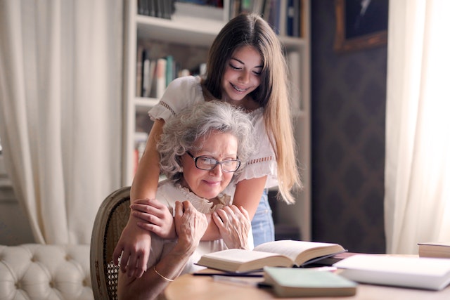 A teenage girl standing behind her grandma who is sitting and hugging her. They are both looking at the books on the table and smiling. (Grandma telling her granddaughter about her memories)