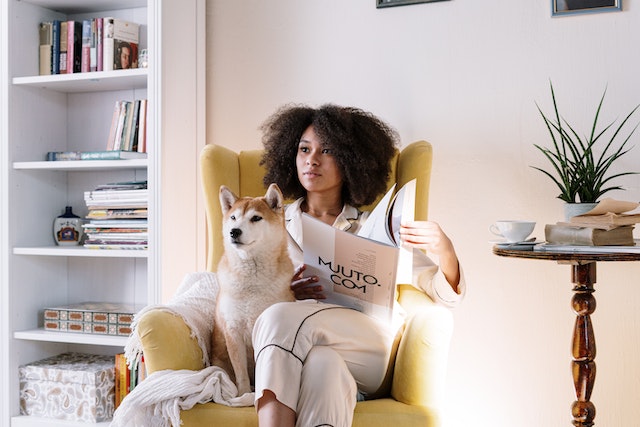 A girl sitting in a yellow chair, reading a newspaper with a dog sitting next to her.(How would you start a conversation online with this girl?)