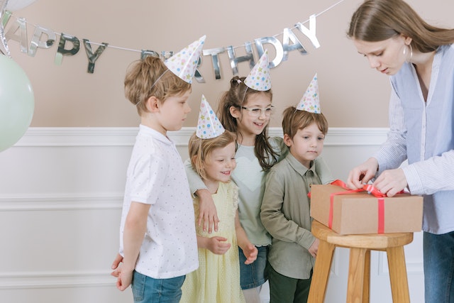A woman unpacks a present in front of the four children with birthday caps on.(Practice saying, "I bought gifts for them"!)
