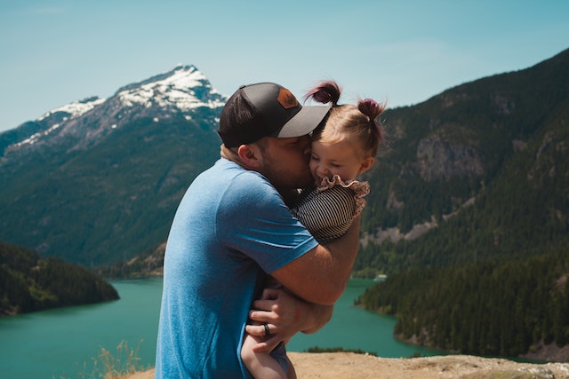 A father holding and kissing his little daughter in front of a river and mountains.
(Serbian teacher says, "Ovaj čovek je sjajan tata"!)