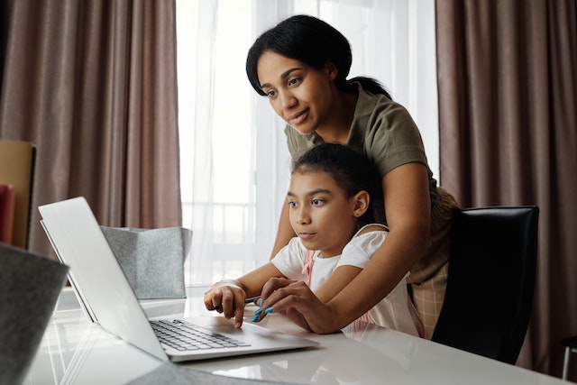 A mom standing above her daughter and helping her type something on a laptop.
(For teaching someone something, use the verb učiti!)