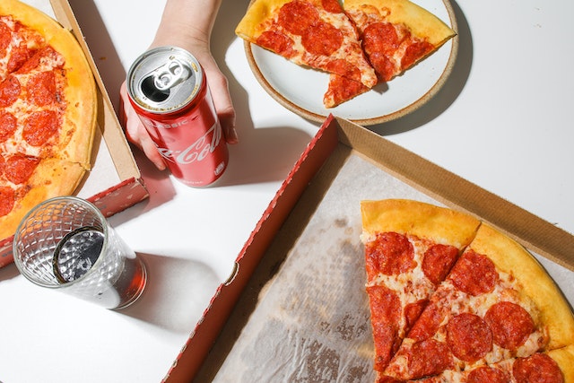 Two opened pizza boxes with pizzas in them, a plate with two slices of pizza, a hand holding a can of Coca-Cola, and a glass of Coca-Cola.(A Serbian tutor won't tell you they chose the "za poneti" option!)