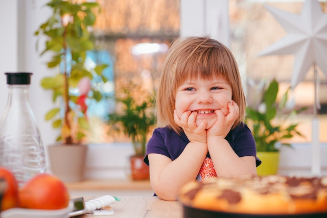 A girl sitting at the table smiling and holding her face. There's a pie, oranges, and an empty bottle of juice in front of her.(It's easy to talk about one child in Serbian!)