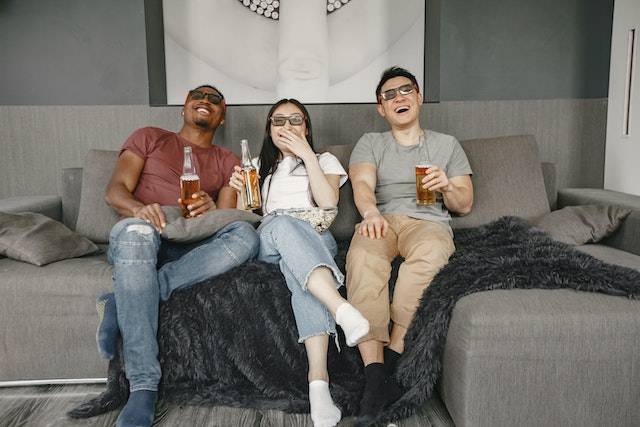 Two boys and a girl sitting on a couch, laughing and watching a movie. They're drinking beer and she's holding a bowl of popcorn.(Learning Serbian with popcorn is fun!)