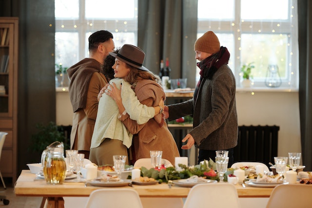 Four people standing around the table with a holiday feast. Two women are hugging, and the men are shaking hands.(Learning Serbian Christmas traditions: say "Hristos se rodi" to wish merry Christmas in Serbia!)