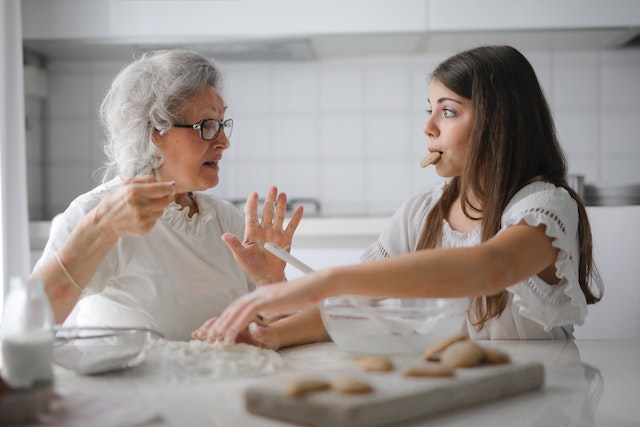 A grandmother and a granddaughter are baking cookies. The grandmother is explaining something, while the granddaughter is eating a cookie.
(Is there a better way of honoring the Serbian language than learning from your grandparents)