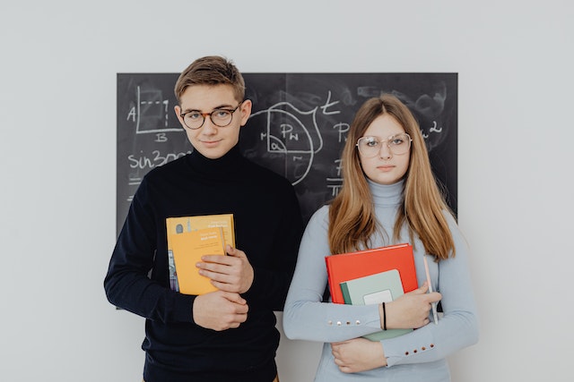 Two high school students (a boy in a black sweater and a girl in a blue sweater) standing in front of the blackboard holding books.
(Learning Serbian false friends: gimnazija is a type of high school!)