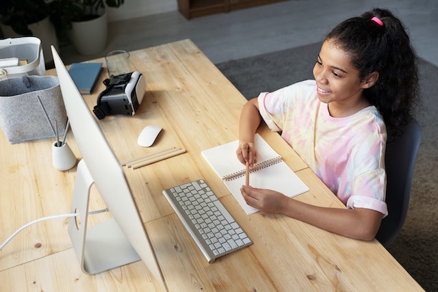 A girl sitting at the wooden table looking at her computer. In front of her is a notebook and she's holding a pencil.
(How long have you been learning Serbian?)