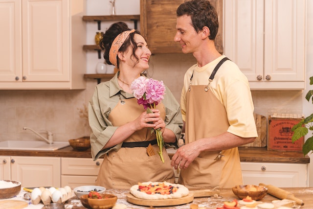 A man giving flowers to a woman in the kitchen. They both wear aprons. (Learning Serbian through life: Giving flowers to your significant other is nice, but doing your share of the chores is better!)