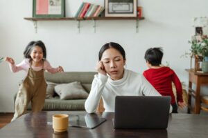 A tired woman is sitting in front of her computer at home while her two little children run and play around her. (A Serbian online course might not be the best idea if you have too many distractions.)