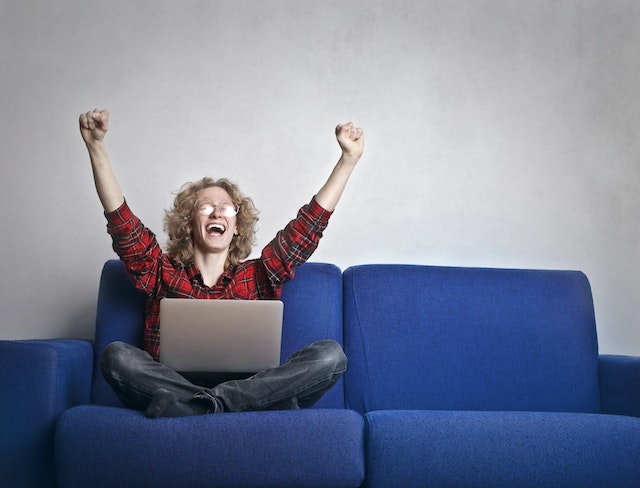 A woman in a red shirt and jeans sitting on a blue couch with a laptop in her lap. She's triumphantly holding both hands in the air and has a wide smile.