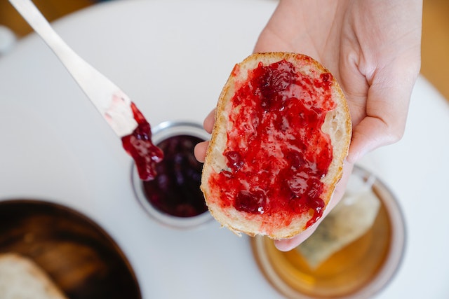 A hand holding a piece of bread with jelly and a wooden spoon in the jar of jelly.