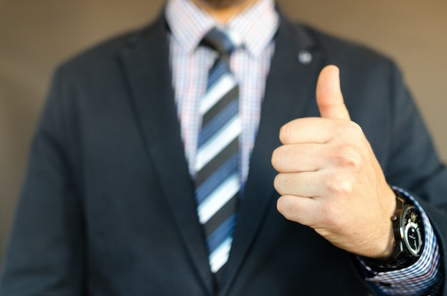 A man showing a thumb up. He's wearing a suit and a watch. We can only see his torso, without head and legs.
