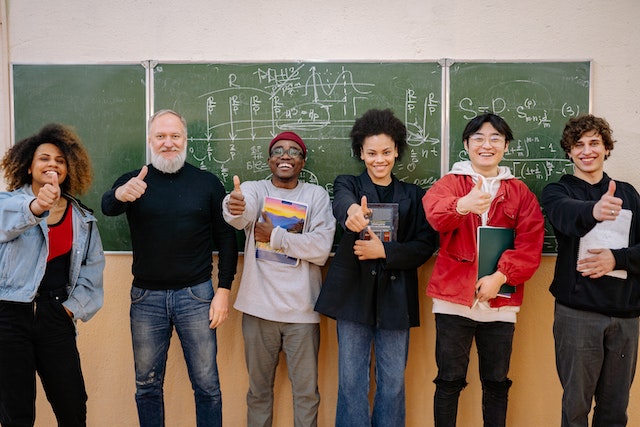 Six people from different backgrounds people standing in front of the blackboard, holding notebooks, smiling and showing thumbs up.