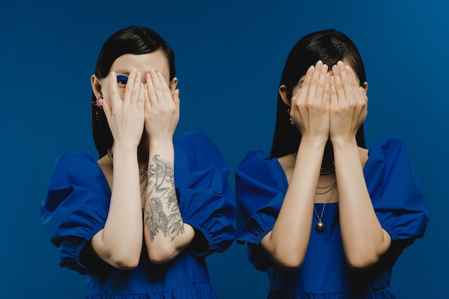 Two black-haired women in the same blue shirts are in front of the blue background. They both covered their faces with their arms, but only one is peeking.
(The Serbian cases Dative and Locative seem the same, but are they?)