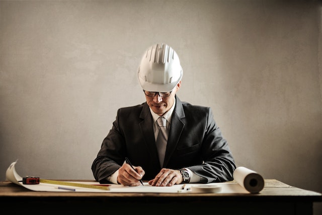 A man in a suit and with a helmet on his head drawing at the desk with a meter and a long piece of paper.)