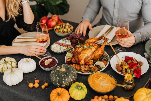 A table full of food (roasted chicken, pumpkins, apples, and vegetables). A man and a woman are sitting at the table, holding two glasses of wine.