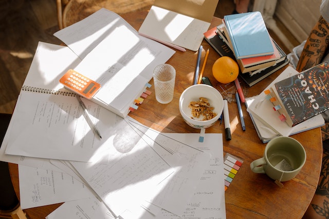 A wooden table with many open notebooks, a calculator, a cup of tea, cereal, books, and marker.