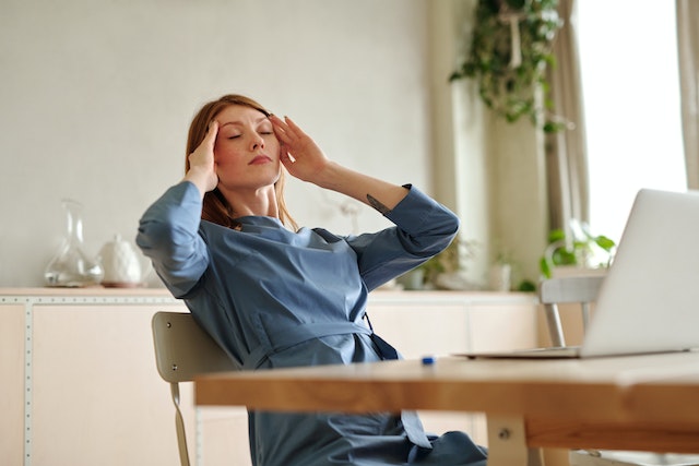 A woman in a blue dress sitting in front of her laptop and holding her head in pain.