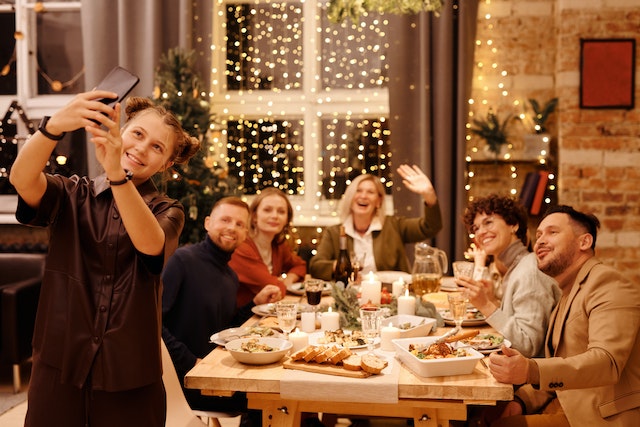 A happy family of 6 adult people sitting at the table, having dinner, and smiling to the camera. A girl is standing in front of the table taking a selfie with all of them.